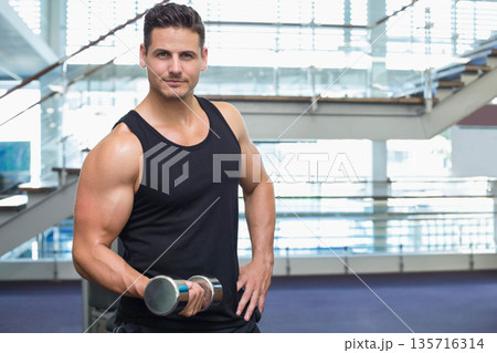 Muscular man 30s wearing black tank and dark shorts holding chrome dumbbell posing in gym mezzanine 135716314