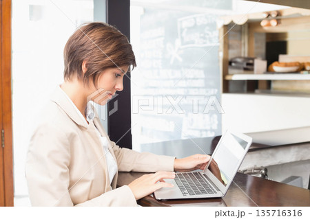 Woman in 20s-30s sitting at cafe counter, typing on laptop by chalkboard menu, wearing beige blazer 135716316