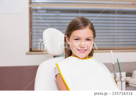 School-age girl sitting in dental chair wearing white bib with yellow trim, dental tray nearby School-age girl sitting in dental chair wearing white bib with yellow trim, dental tray nearby 135716318