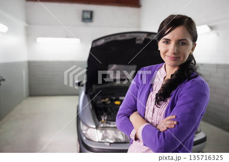 Adult woman standing arms crossed in garage bay by silver sedan with open hood, copy space Adult woman standing arms crossed in garage bay by silver sedan with open hood, copy space 135716325