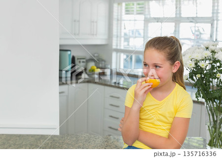 Female child sitting on kitchen counter wearing yellow tee holding half orange biting, copy space Female child sitting on kitchen counter wearing yellow tee holding half orange biting, copy space 135716336