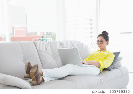 Adult Asian woman reclining on sofa in living room wearing yellow blouse using laptop, copy space Adult Asian woman reclining on sofa in living room wearing yellow blouse using laptop, copy space 135716350