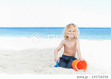 Child boy sitting shirtless on white sand by ocean, playing with orange bucket and blue shovel 135716356