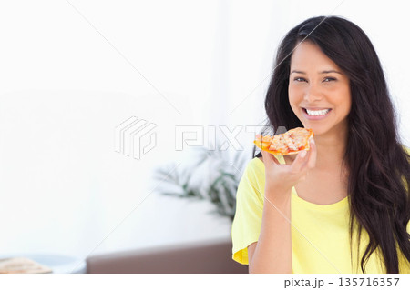 Woman holding pizza slice, smiling toward camera in bright living room with yellow tee, copy space Woman holding pizza slice, smiling toward camera in bright living room with yellow tee, copy space 135716357