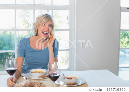 Mature woman sitting at dining table holding phone, wine, soup and loaf, wearing blue top Mature woman sitting at dining table holding phone, wine, soup and loaf, wearing blue top 135716362
