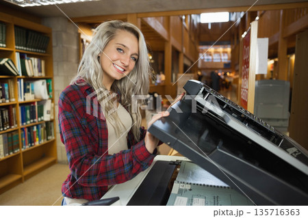 Adult woman in plaid shirt lifting copier lid and placing spiral notebook at library near shelves 135716363