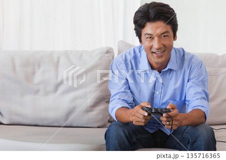 Mid adult Asian man wearing light blue shirt and dark jeans, playing wired controller on sofa 135716365