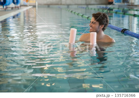 Adult man standing chest-deep at pool facility holding two pink foam noodles, shirtless, copy space 135716366