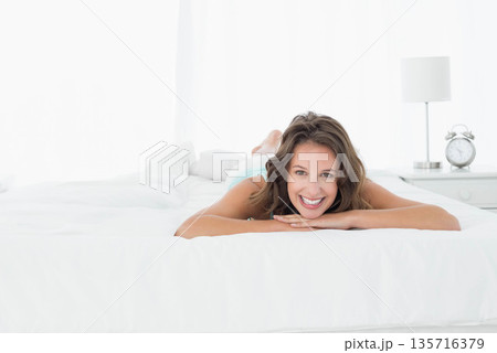 Adult woman propped on arms smiling at camera on white bed wearing blue tank, nightstand clock 135716379