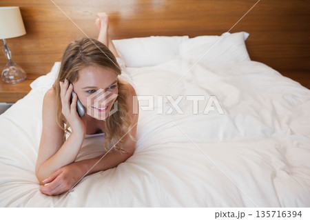 Woman lying on stomach on large bed wearing light camisole holding silver smartphone to ear smiling Woman lying on stomach on large bed wearing light camisole holding silver smartphone to ear smiling 135716394