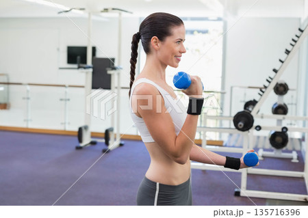 Female exerciser performing dumbbell curls in gym wearing white sports bra and gray leggings 135716396