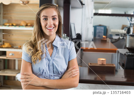 Female barista standing at counter in light blue blouse, showing wooden shelves and coffee grinder Female barista standing at counter in light blue blouse, showing wooden shelves and coffee grinder 135716403