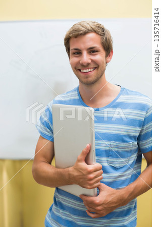 Man wearing blue striped tee standing in classroom, smiling and holding slim laptop near whiteboard 135716414