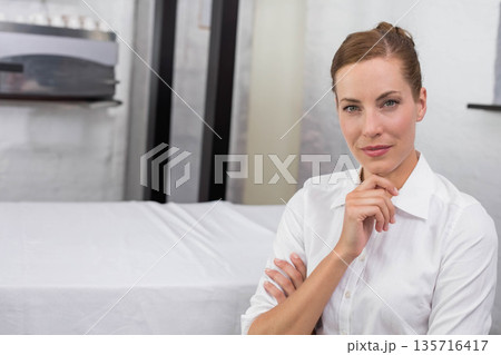 Mid-adult woman standing hand under chin in white shirt in treatment room near table, copy space Mid-adult woman standing hand under chin in white shirt in treatment room near table, copy space 135716417