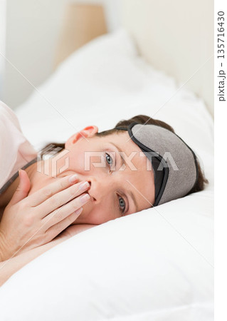 Woman lying on bed with white pillow and bedside lamp, wearing gray sleep mask, covering mouth 135716430