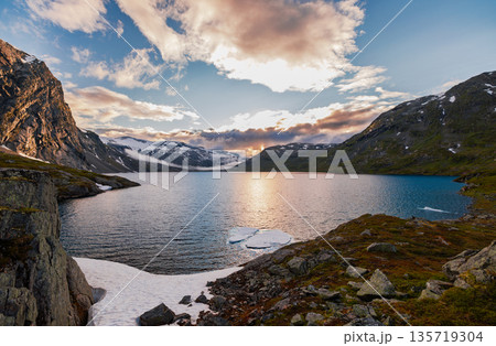 Serene Norwegian mountain lake with melting ice, snow patches and dramatic sunset clouds reflecting in calm turquoise water. Serene Norwegian mountain lake with melting ice, snow patches and dramatic sunset clouds reflecting in calm turquoise water. 135719304