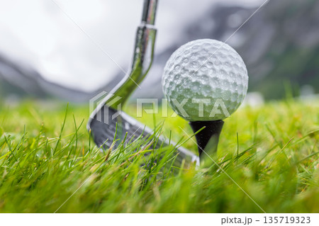 Golf club and ball on wet green field with mountains in background. 135719323