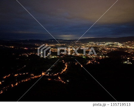 nighttime road scene, serpentine vehicle lights illuminate distant suburban outskirts with soft silhouettes 135719513