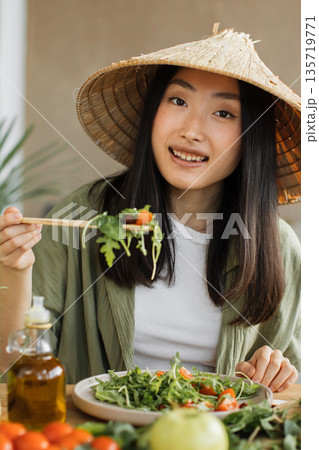 A young Asian woman wearing a conical hat enjoys a healthy salad with chopsticks, surrounded by fresh ingredients and olive oil A young Asian woman wearing a conical hat enjoys a healthy salad with chopsticks, surrounded by fresh ingredients and olive oil 135719771