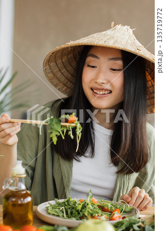 A young Asian woman wearing a conical hat enjoys a fresh salad with chopsticks, surrounded by healthy ingredients and olive oil 135719772