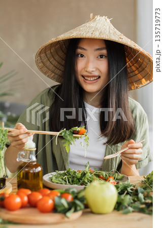 A young Asian woman wearing a conical hat enjoys a healthy salad with fresh vegetables and olive oil 135719773