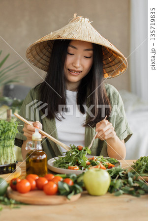 A young Asian woman wearing a conical hat enjoys a fresh salad with chopsticks, surrounded by healthy ingredients 135719835