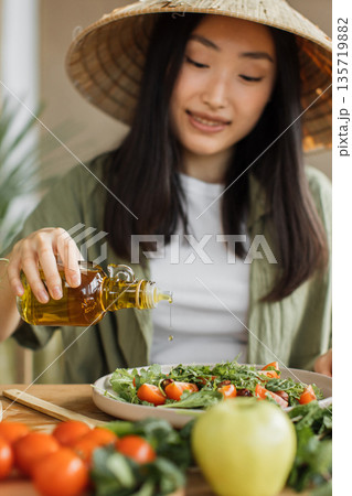 A young woman wearing a conical hat pours olive oil onto a fresh salad with tomatoes and greens 135719882