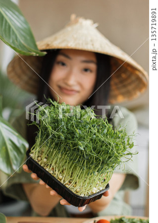 A young woman wearing a conical hat holds a tray of vibrant green microgreens, showcasing healthy, home-grown produce 135719891