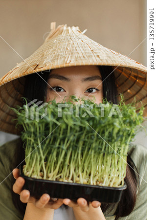 A young woman wearing a conical hat holds a tray of fresh microgreens in front of her face, showcasing healthy eating and sustainable agriculture A young woman wearing a conical hat holds a tray of fresh microgreens in front of her face, showcasing healthy eating and sustainable agriculture 135719991
