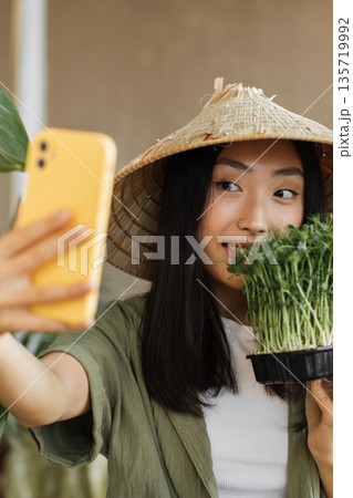 A young Asian woman wearing a conical hat takes a selfie with a tray of fresh microgreens in her hand A young Asian woman wearing a conical hat takes a selfie with a tray of fresh microgreens in her hand 135719992