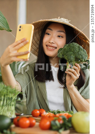 A young Asian woman wearing a conical hat takes a selfie with a broccoli floret while surrounded by fresh vegetables on a wooden table 135719994