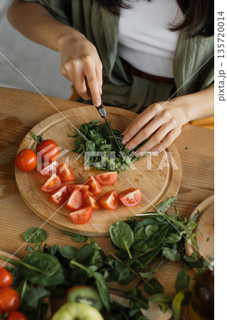 A person is preparing a healthy meal by chopping fresh greens and tomatoes on a wooden cutting board, surrounded by more vegetables A person is preparing a healthy meal by chopping fresh greens and tomatoes on a wooden cutting board, surrounded by more vegetables 135720014
