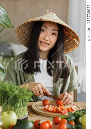 A young Asian woman wearing a conical hat is preparing fresh vegetables for a meal in a bright, natural setting A young Asian woman wearing a conical hat is preparing fresh vegetables for a meal in a bright, natural setting 135720035