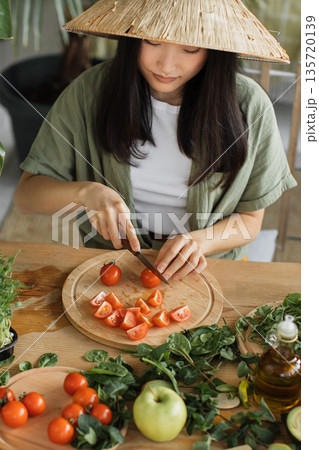 A woman wearing a conical hat prepares fresh ingredients for a healthy meal, chopping tomatoes on a wooden cutting board surrounded by greens and an apple 135720139