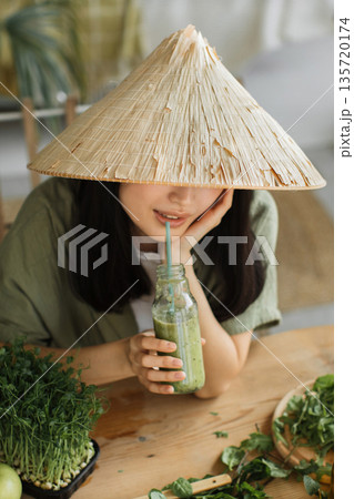 A young woman wearing a conical hat sips a healthy green smoothie from a glass bottle with a straw, surrounded by fresh ingredients on a wooden table 135720174