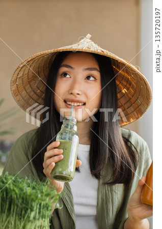 A young Asian woman wearing a conical hat drinks a healthy green smoothie from a glass bottle with a straw, surrounded by fresh microgreens A young Asian woman wearing a conical hat drinks a healthy green smoothie from a glass bottle with a straw, surrounded by fresh microgreens 135720197