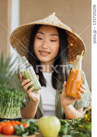 A young Asian woman wearing a conical hat holds two bottles of fresh juice, one green and one orange, surrounded by microgreens, tomatoes, and an apple 135720206
