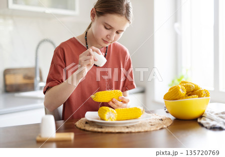 Young Girl Salting Boiling Corn Cobs For Eating In The Kitchen Young Girl Salting Boiling Corn Cobs For Eating In The Kitchen 135720769