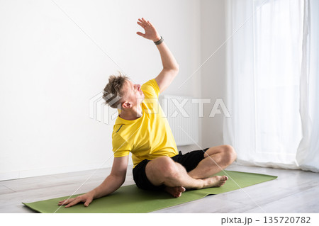 Sporty Young Man Practices A Yoga Routine And Does Core Stretching While Sitting On The Floor At Home 135720782