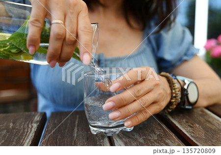 Girl Sitting In An Open-Air Cafe Pouring Fresh Water With Mint And Lemon Into A Glass Girl Sitting In An Open-Air Cafe Pouring Fresh Water With Mint And Lemon Into A Glass 135720809
