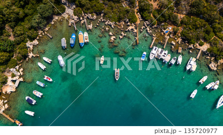 Aerial view of small boats and yachts anchored in a turquoise bay with clear water, wooden piers, rocky shoreline, and lush green trees. Summer seaside marina from drone. Aerial view of small boats and yachts anchored in a turquoise bay with clear water, wooden piers, rocky shoreline, and lush green trees. Summer seaside marina from drone. 135720977