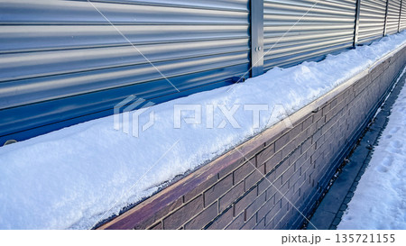 A thick layer of fresh white snow lying on a brown brick wall next to blue metal roller shutters. A thick layer of fresh white snow lying on a brown brick wall next to blue metal roller shutters. 135721155