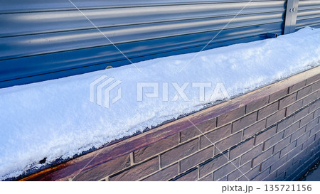 A thick layer of fresh white snow lying on a brown brick wall next to blue metal roller shutters. A thick layer of fresh white snow lying on a brown brick wall next to blue metal roller shutters. 135721156