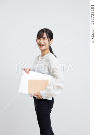 白背景の前でノートパソコンとノートを持ってカメラ目線で微笑む若い女性 白背景の前でノートパソコンとノートを持ってカメラ目線で微笑む若い女性 135722101