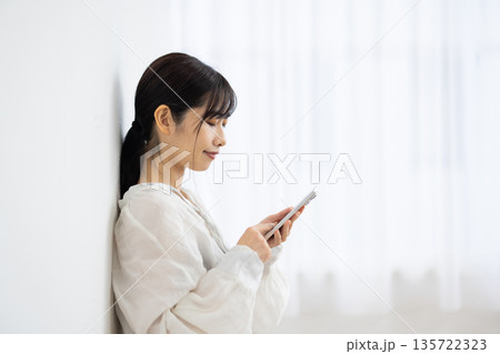 白い壁にもたれてスマートフォンを操作する若い女性の横顔 白い壁にもたれてスマートフォンを操作する若い女性の横顔 135722323