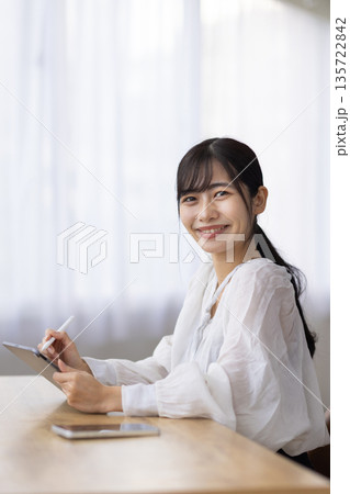 ペンを使ってタブレットを操作しながらカメラ目線で微笑む女性 135722842