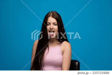 Playful young woman making funny face with tongue out against blue background, humor and carefree emotion concept Playful young woman making funny face with tongue out against blue background, humor and carefree emotion concept 135723890