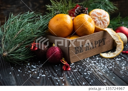 Orange mandarins in wooden box with fir apple, red balls, snowflakes on green fir branch background. Christmas card. Happy new year and merry christmas. High quality photo 135724967