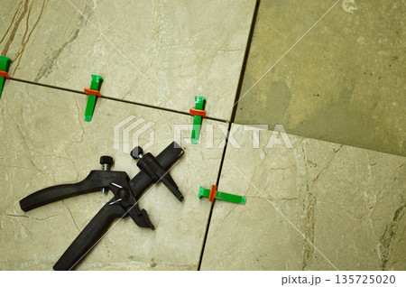 A professional tiler is carefully positioning a ceramic wall tile onto a layer of fresh adhesive, utilizing a lash tile leveling system for a perfectly flat surface. 135725020