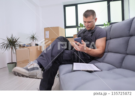 Worker takes a break on a cozy couch while checking phone in a new living space filled with moving boxes and plants Worker takes a break on a cozy couch while checking phone in a new living space filled with moving boxes and plants 135725872